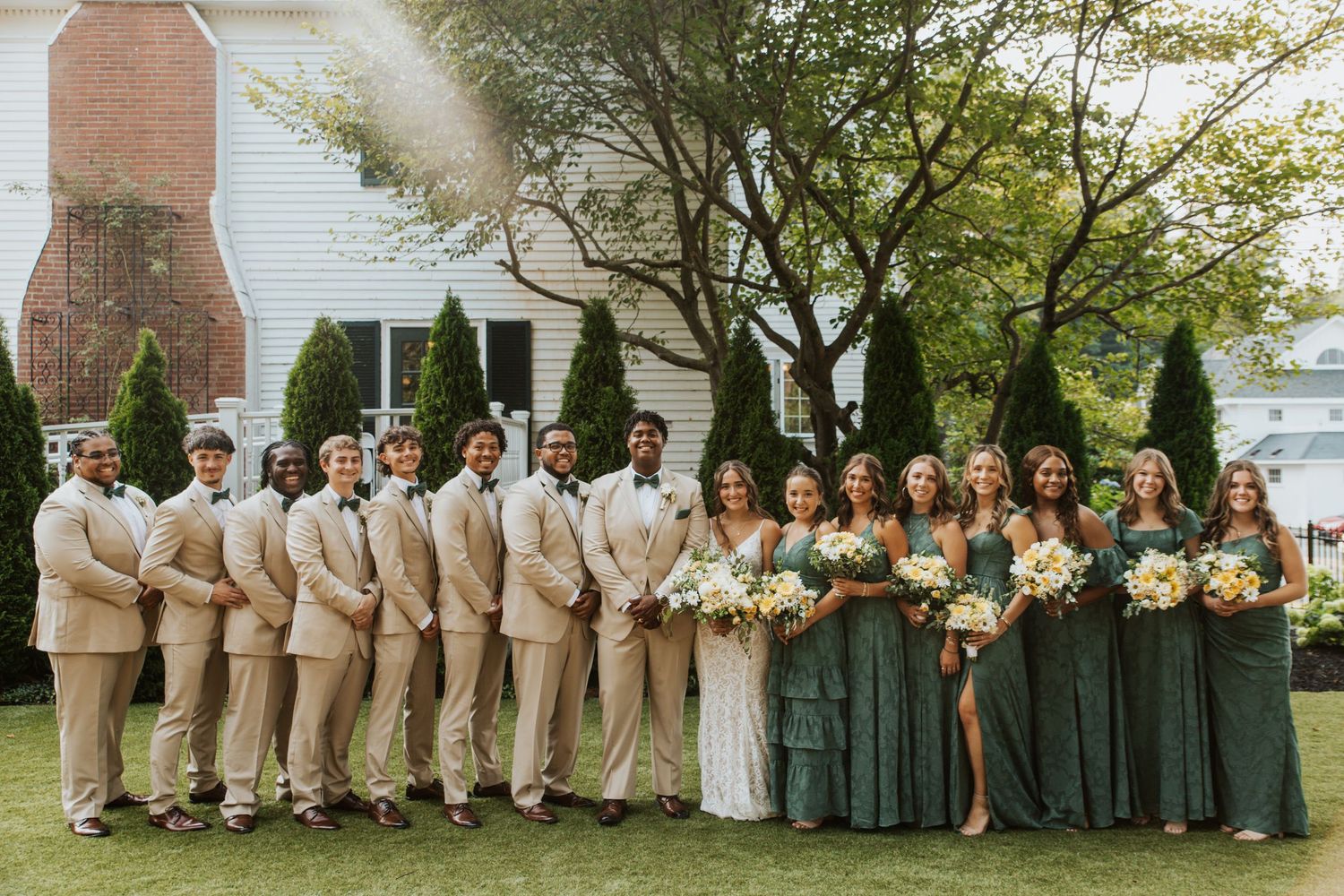 Wedding party poses outdoors, groomsmen in tan suits and bridesmaids in emerald green dresses.