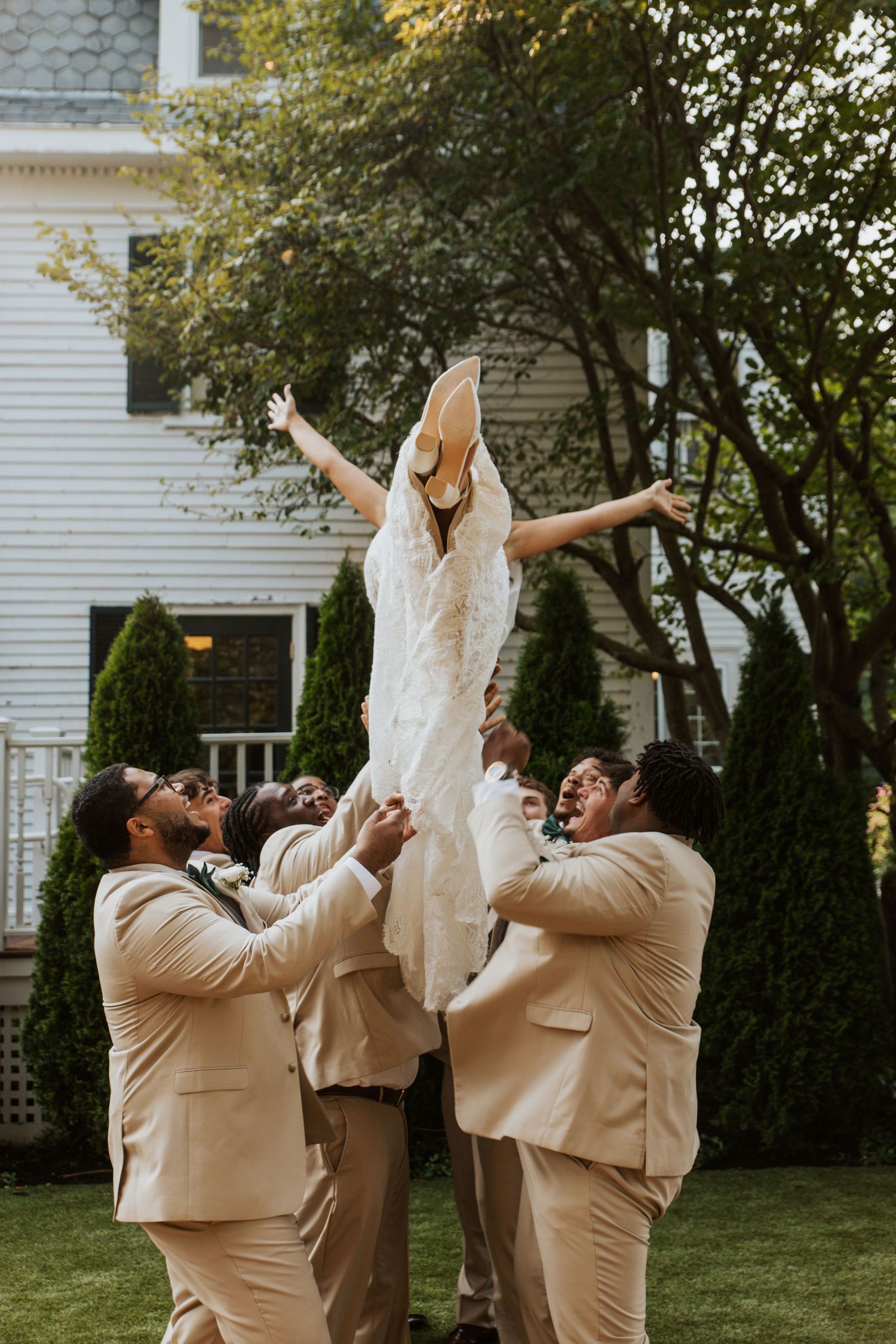 Groomsmen in tan suits joyfully lifting bride in white lace gown outdoors at wedding celebration.