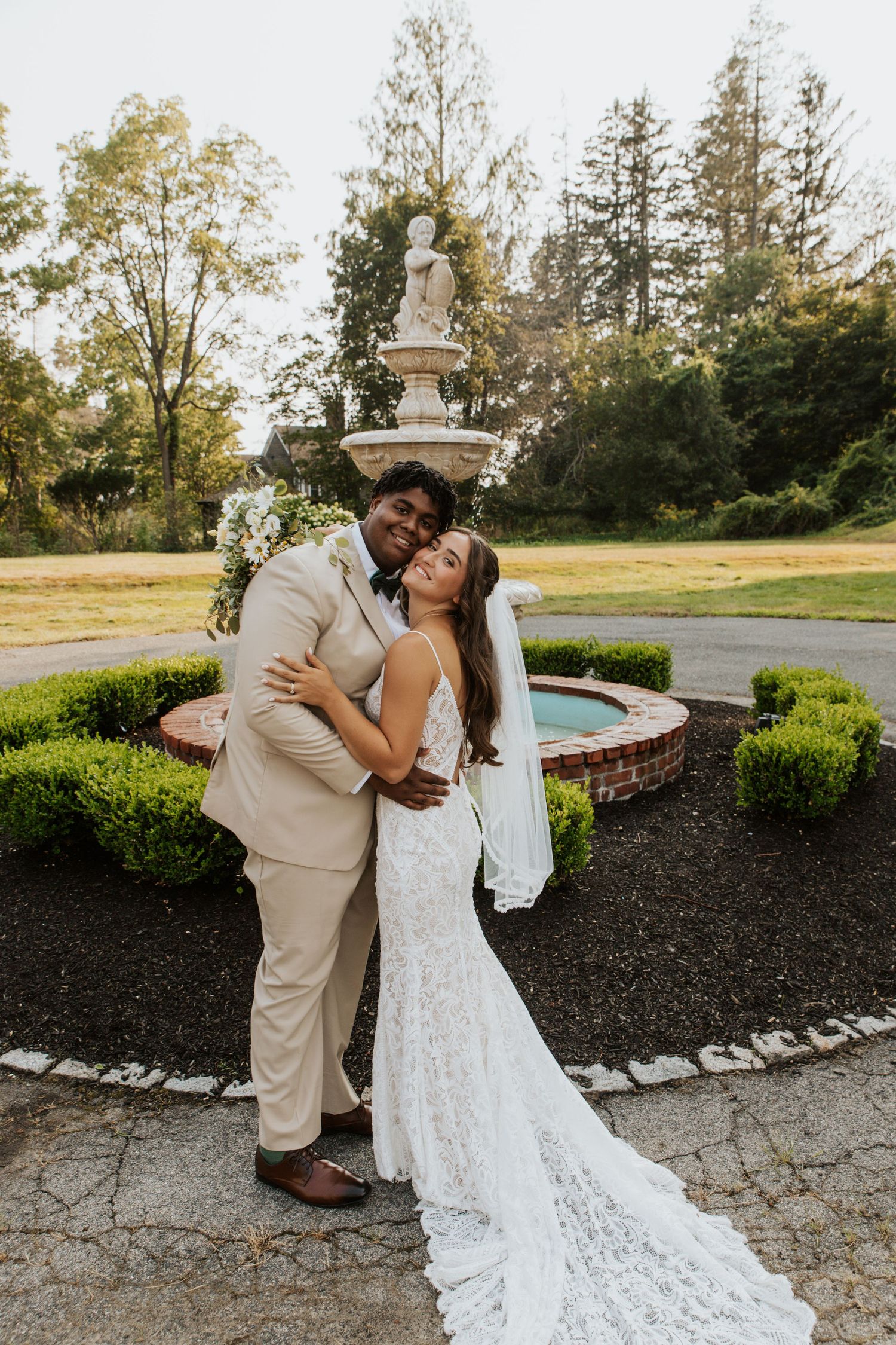 Interracial couple poses by stone fountain, groom in tan suit embracing bride in lace wedding gown.