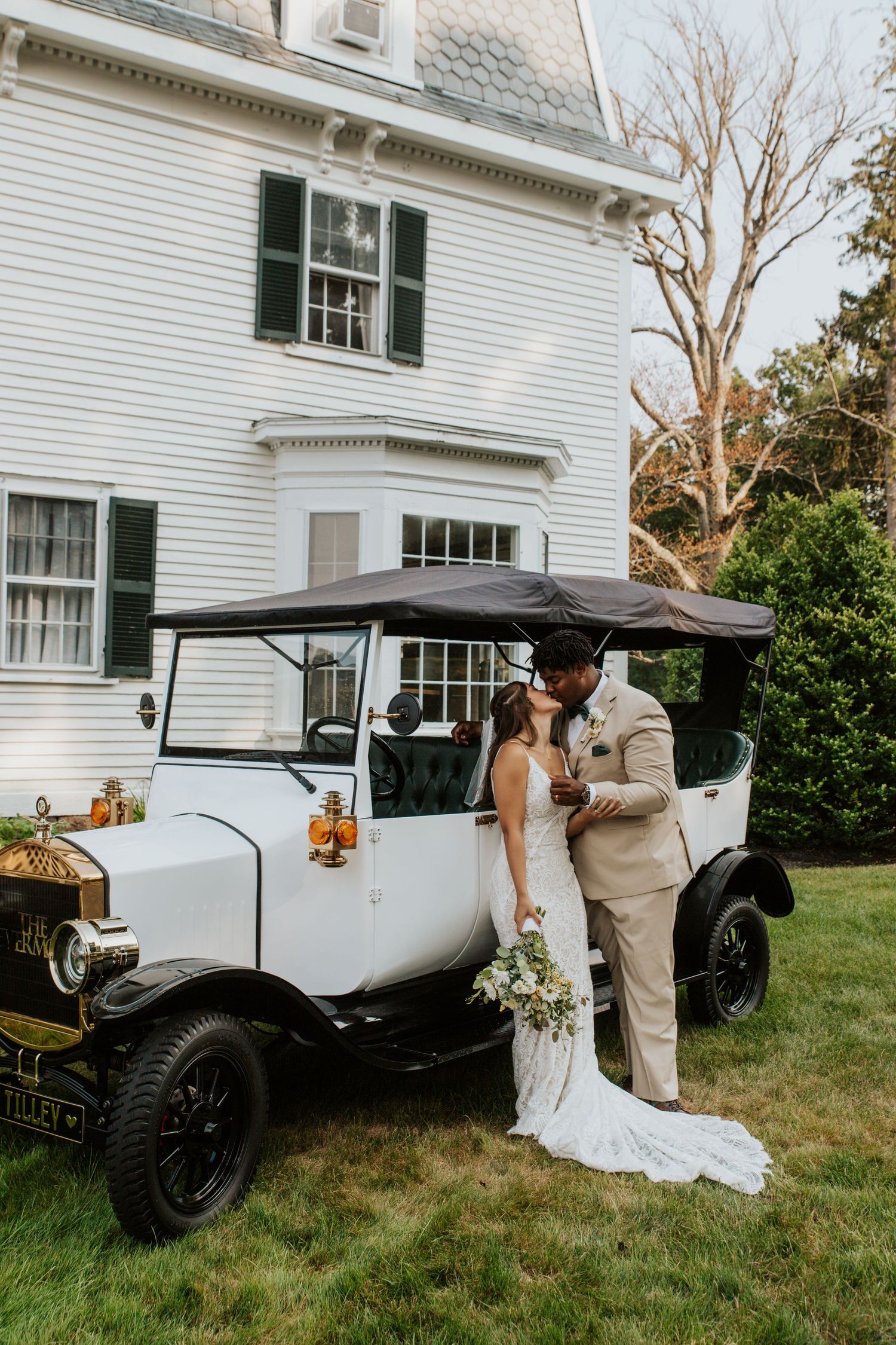 Newlywed couple kissing beside a vintage white antique car outside a classic white estate home.