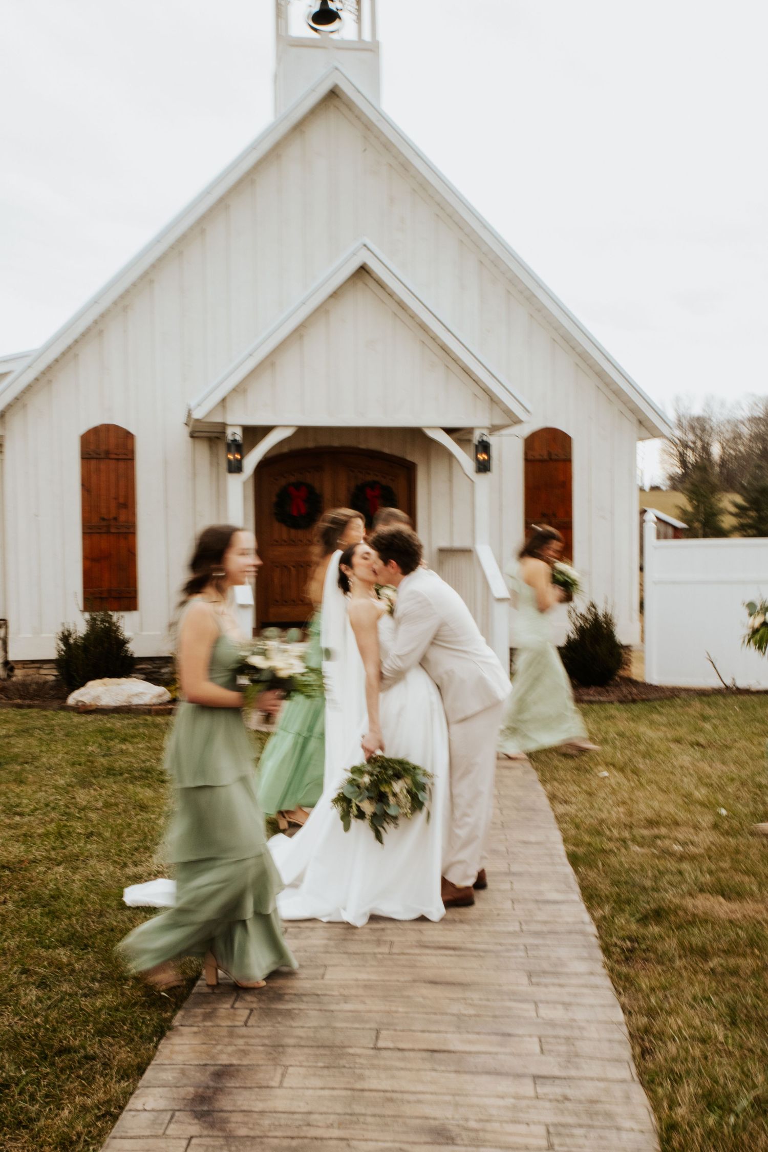 Bride and groom kissing outside a white chapel while bridesmaids in sage green dresses walk past on a brick path.
