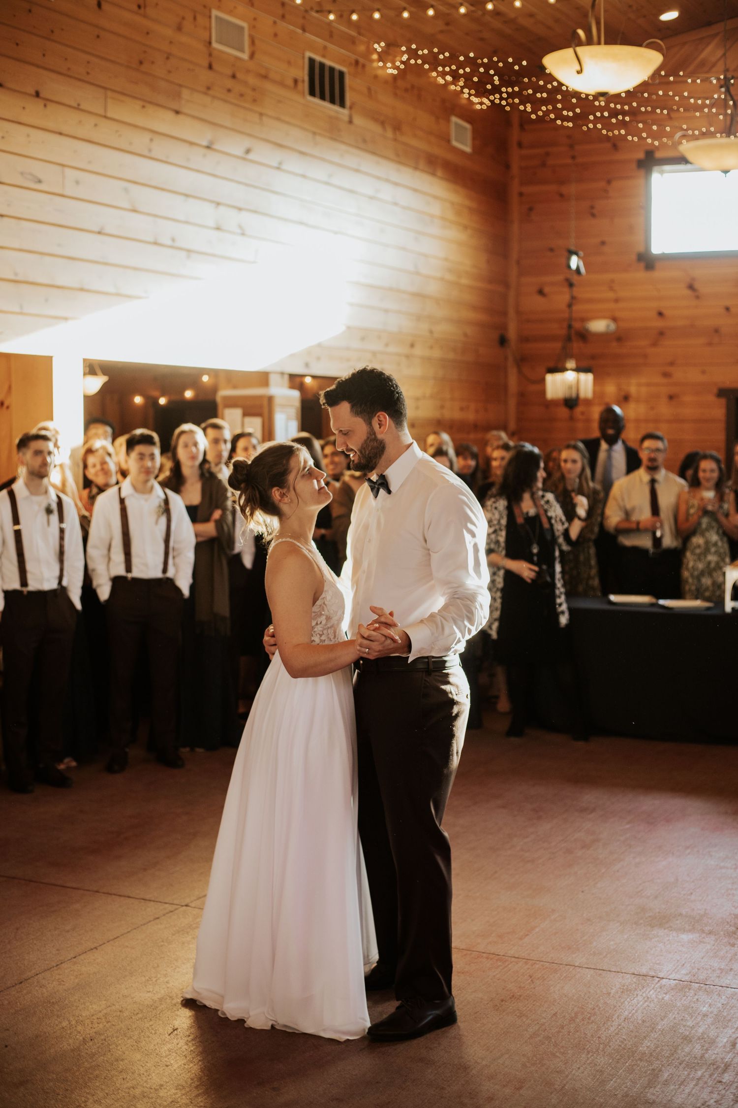 Bride and groom share their first dance in a rustic wooden venue with string lights and guests watching.