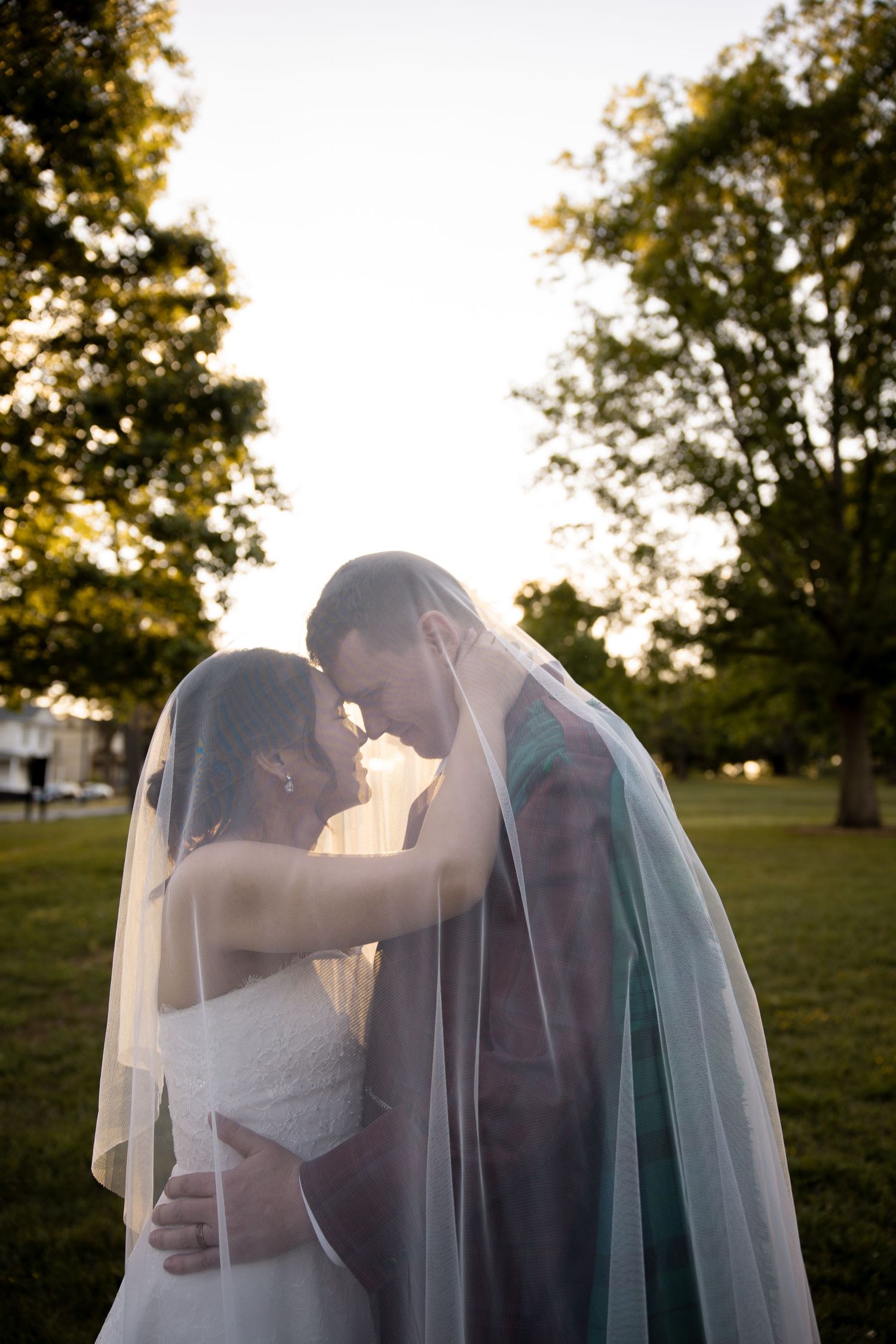 Bride and groom share a romantic moment under a flowing veil outdoors at golden hour.