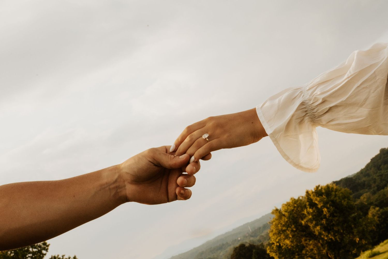 Couple reaching out to hold hands against a scenic cloudy sky, woman wearing an engagement ring.