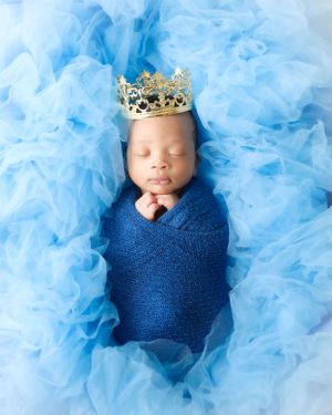 Sleeping newborn wrapped in blue fabric wearing a golden crown against blue tulle background.