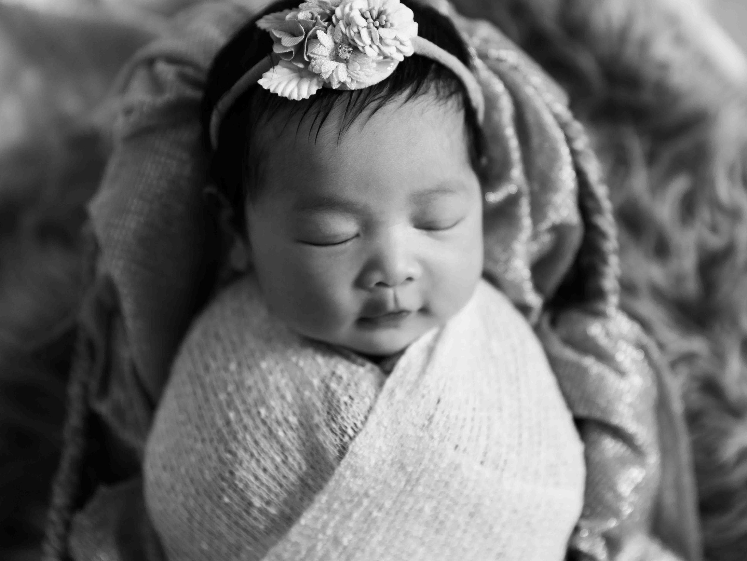 Black and white portrait of sleeping newborn wrapped in soft fabric with floral headband.