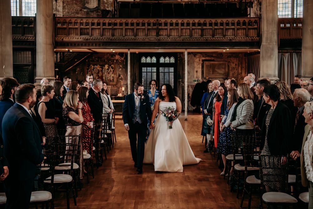 Wedding ceremony in a grand historic hall with wooden beams and guests seated in rows.
