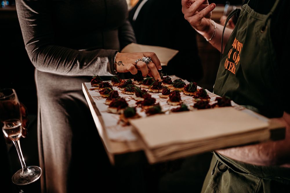 A server holds a wooden board with chocolate desserts while guests reach for wine glasses in a dimly lit setting.