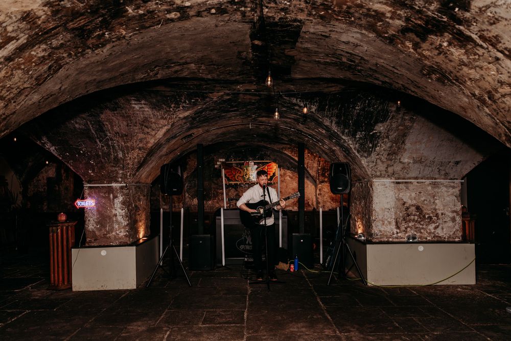 A performer plays music in a rustic underground venue with exposed brick walls and arched ceiling.