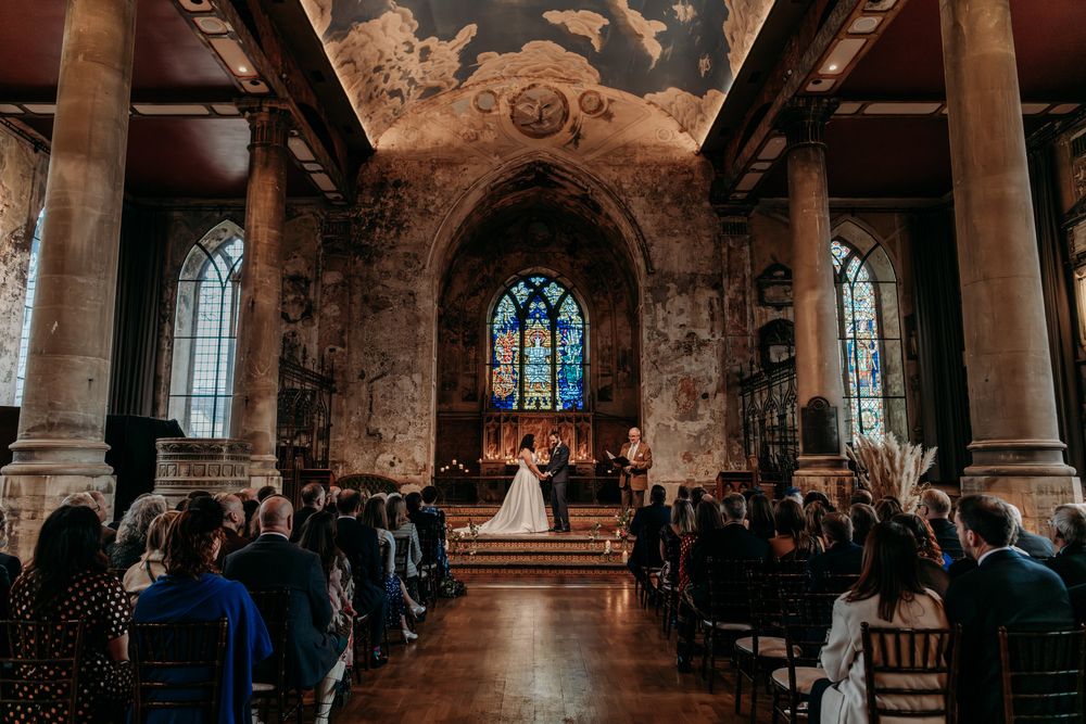 A wedding ceremony takes place in a historic church with stained glass windows and stone columns illuminated by warm light.