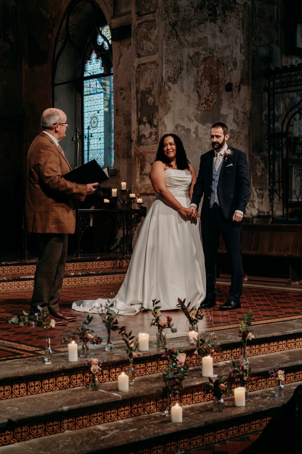 A couple participates in a wedding ceremony inside a historic church with candlelit decorations along the altar steps.