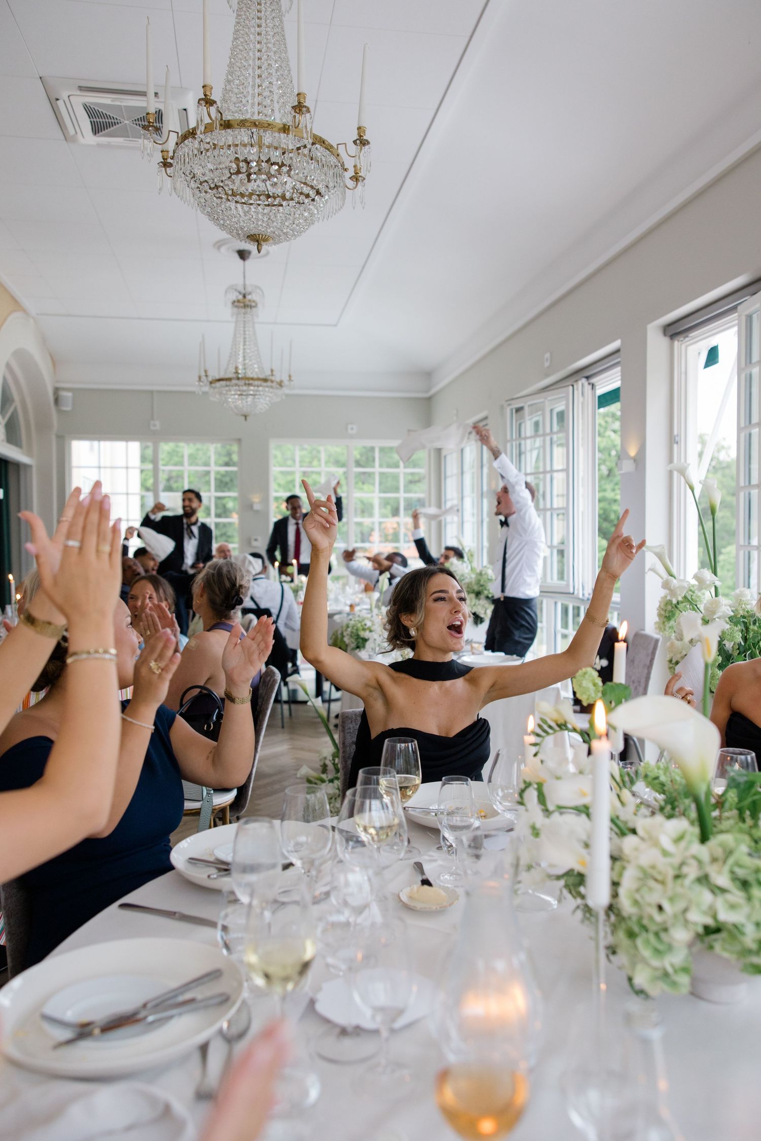 Wedding reception guests wave white napkins at a candlelit table during a celebration in a rustic indoor venue.