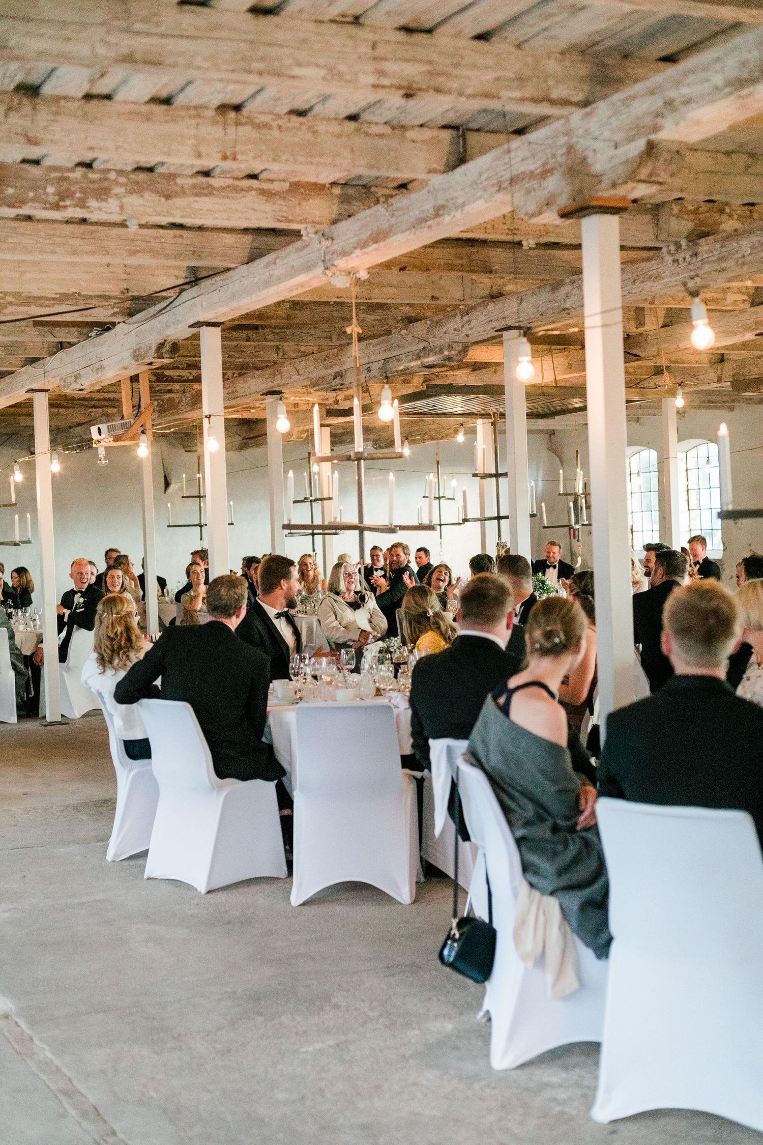 Elegant wedding reception in industrial venue with exposed wooden beams, white chairs and romantic string lighting.