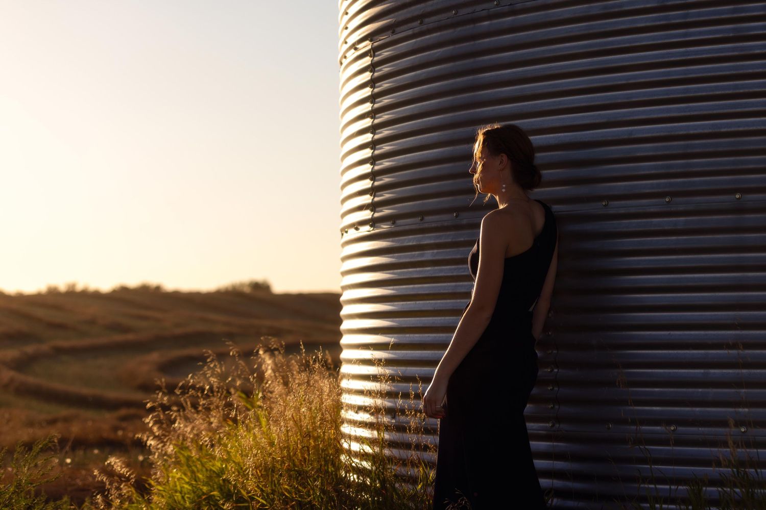 Figure stands in silhouette against metal grain silo during golden sunset.