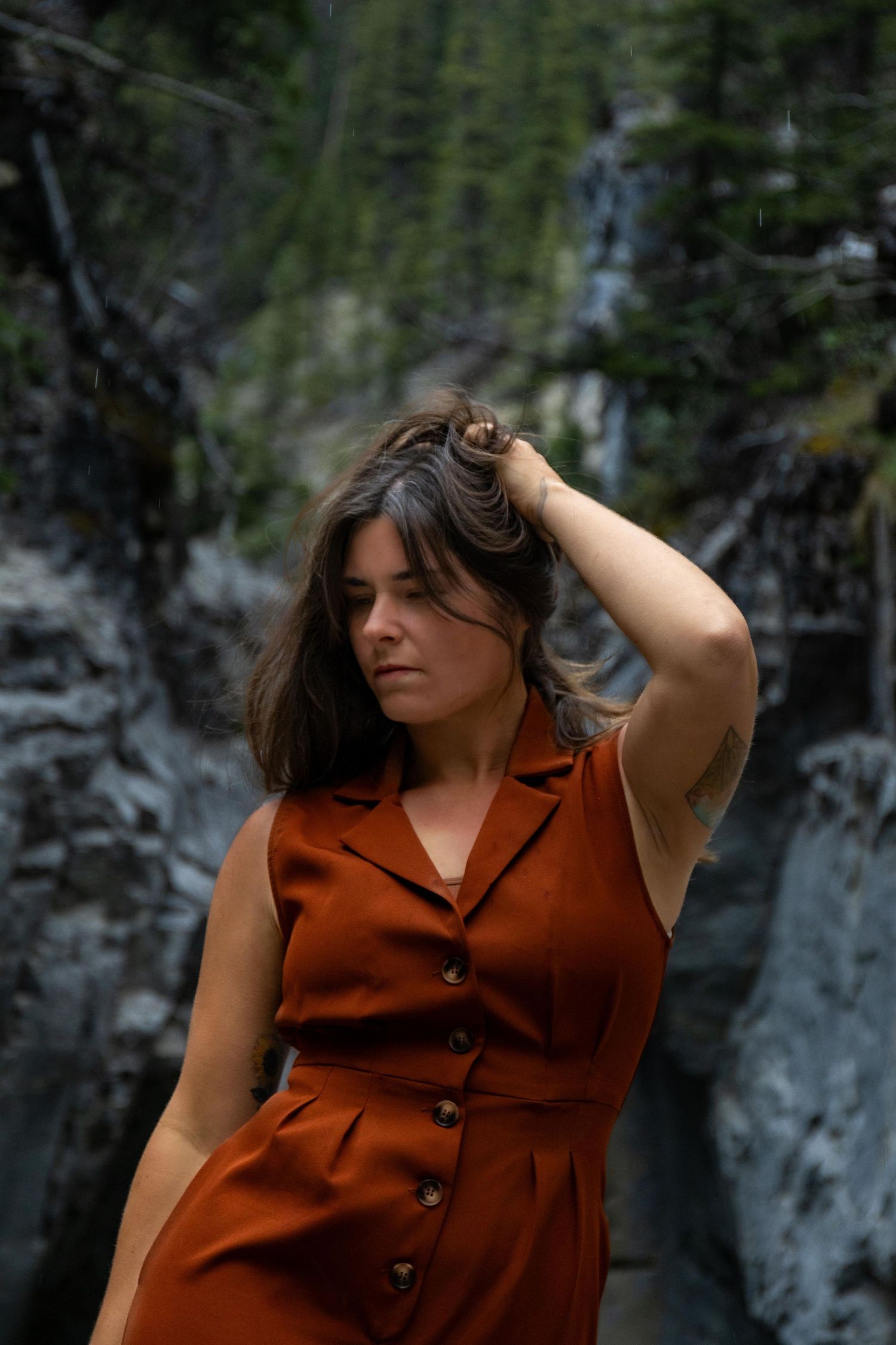 Woman in rust colored jumpsuit poses thoughtfully near a waterfall.