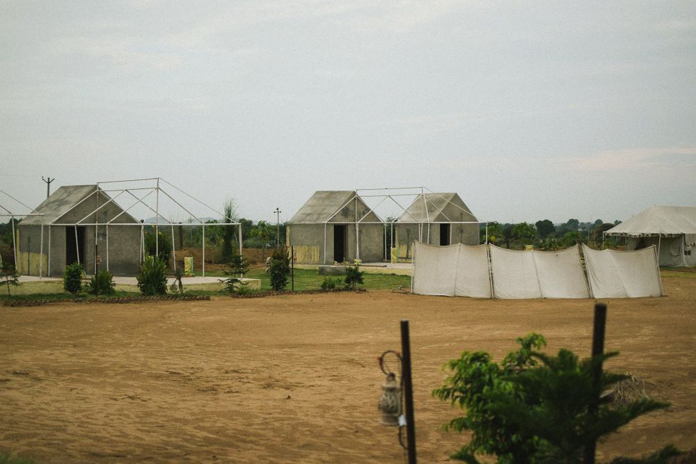 Small cottage houses line a dirt road with white tents in the background under an overcast sky.