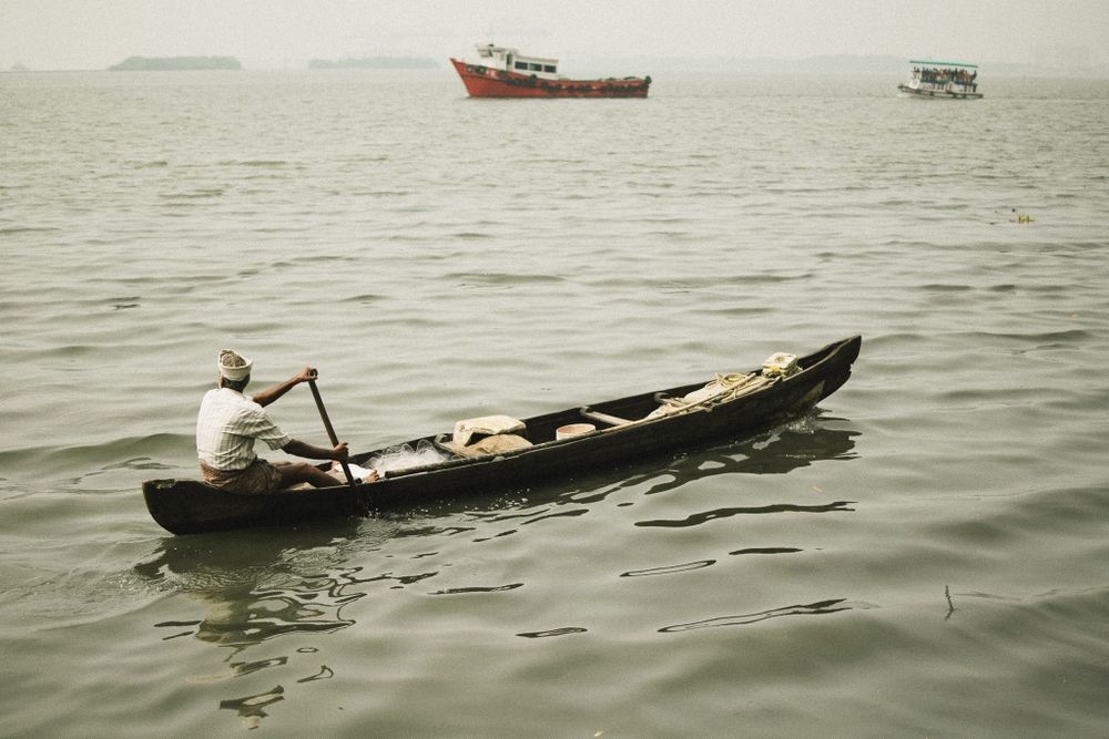 A person rows a traditional wooden canoe across misty waters with larger boats visible in the distance.