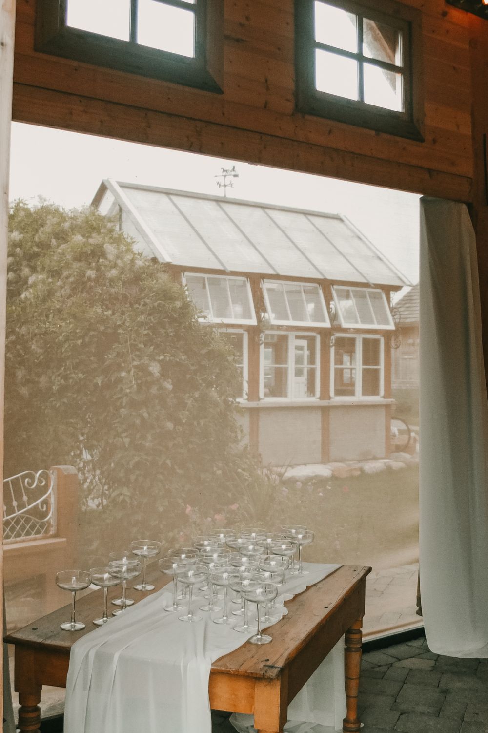 A wooden table with glassware and white linens overlooks a greenhouse through a barn window.
