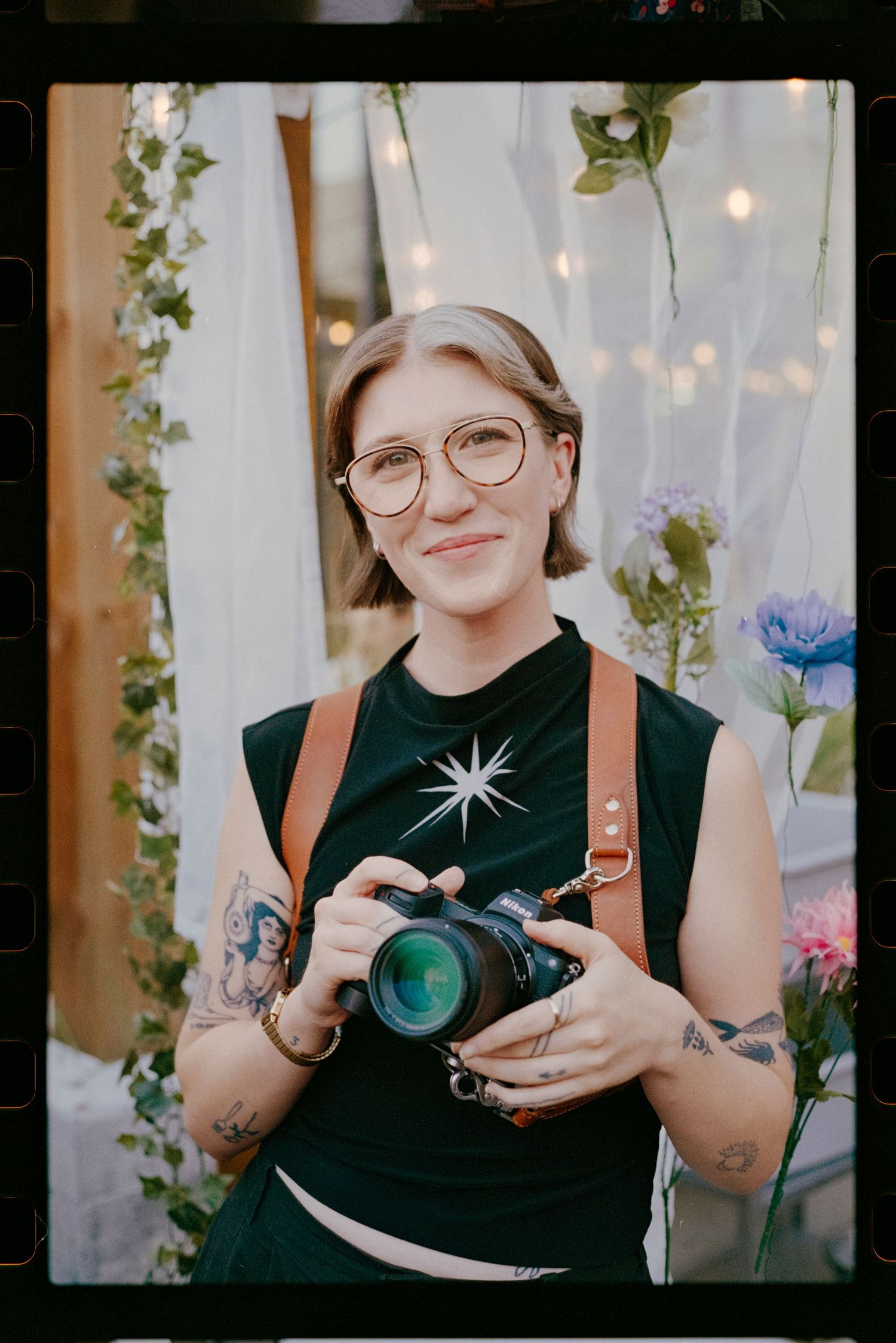 A photographer in a black dress poses with their camera outdoors among tall grasses at golden hour.