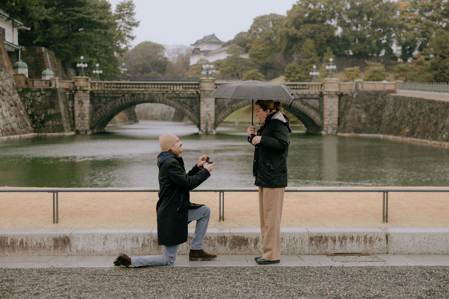 A romantic proposal moment by a historic stone bridge over calm waters on a misty autumn day.