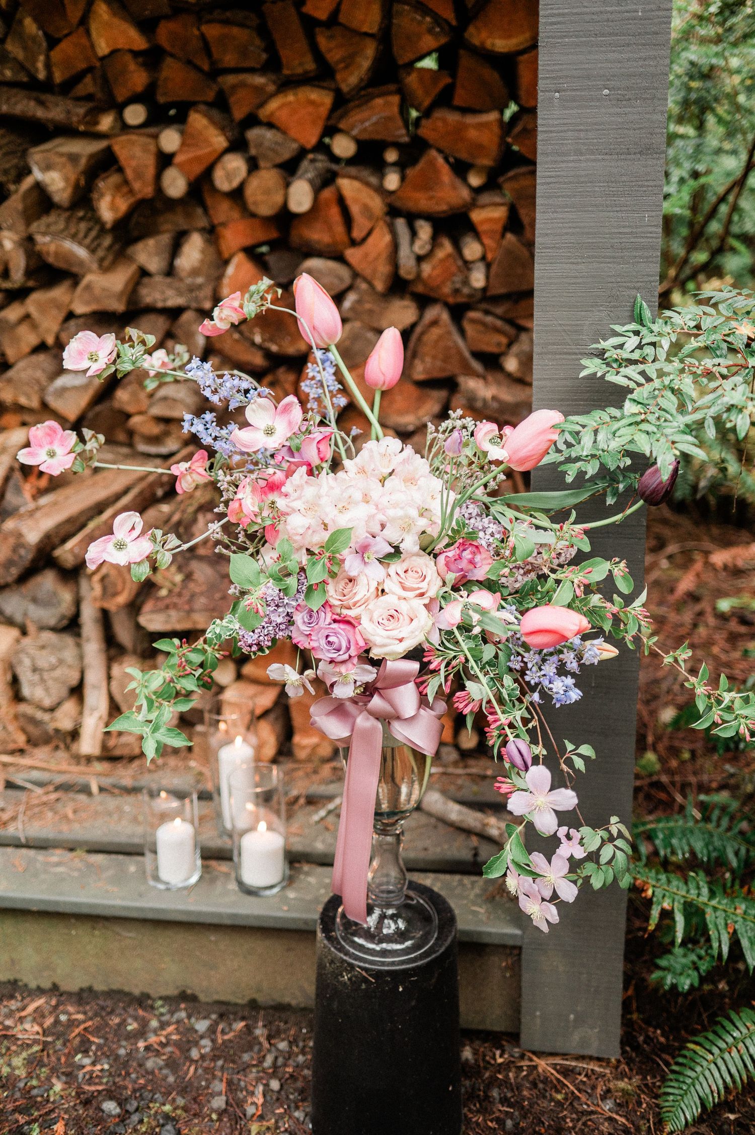 A romantic floral arrangement with pink tulips and roses sits in a black vase against a rustic woodpile backdrop.