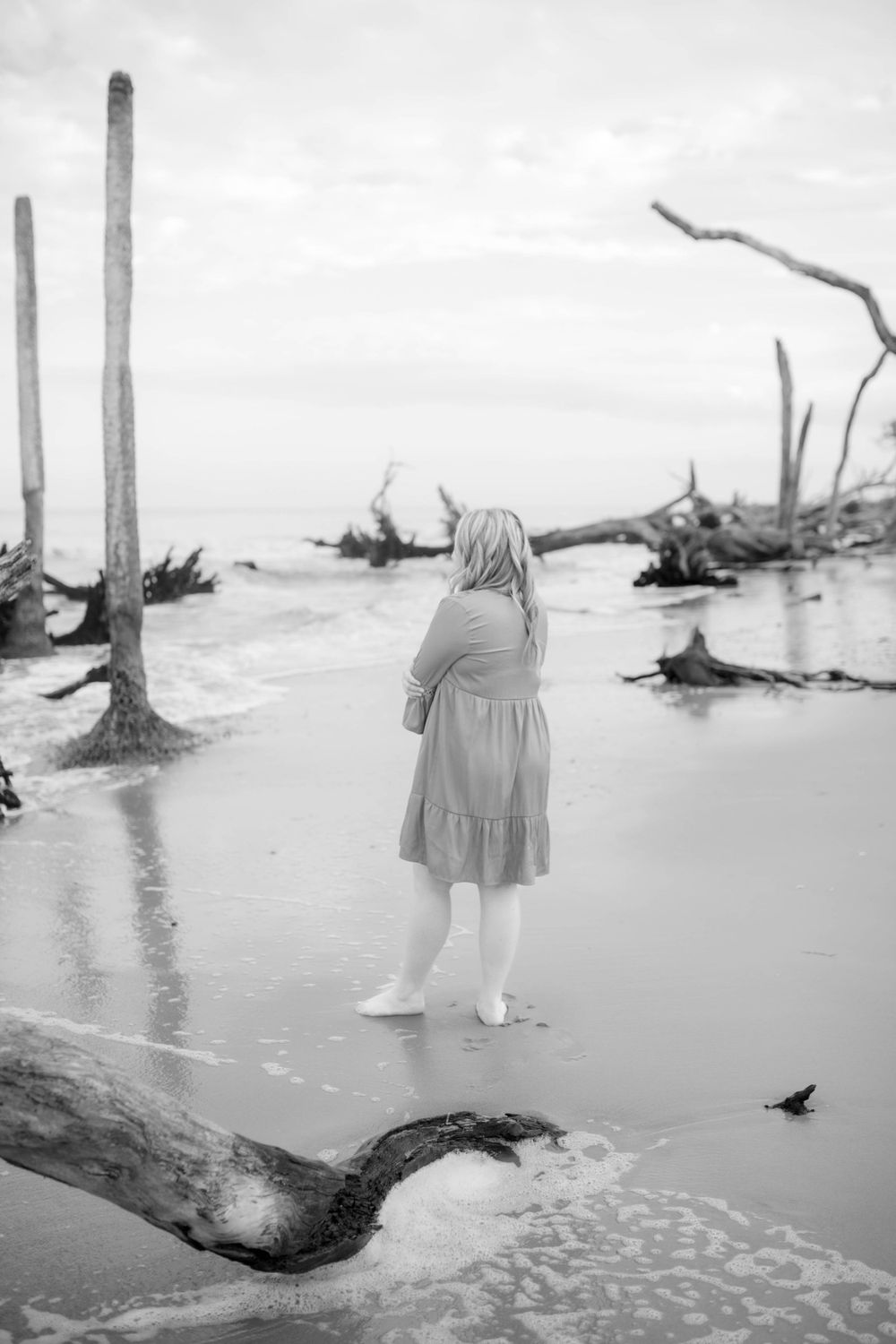 Figure in dress stands among driftwood on misty beach in black and white.