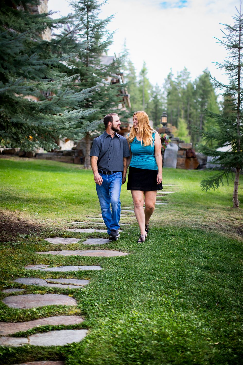 A couple walks along a winding stone path through a green lawn surrounded by evergreen trees.