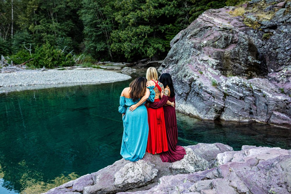 Two people in flowing dresses sit together on rocky outcrop overlooking turquoise mountain lake.