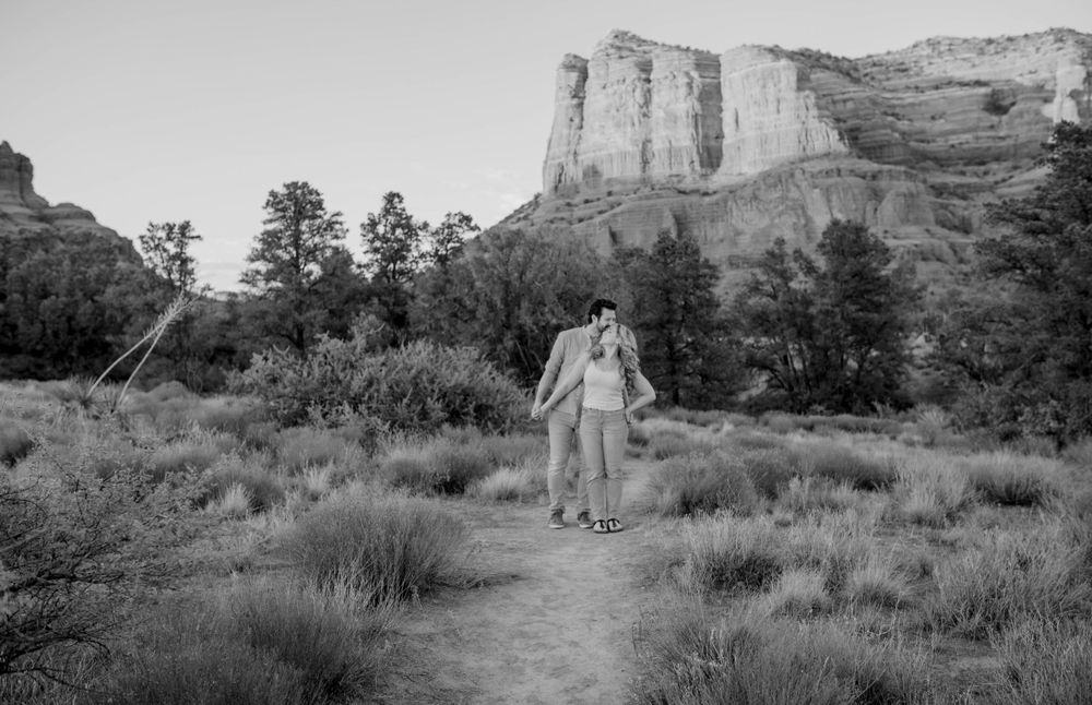 Couple walks along desert trail with majestic red rock formations in background.