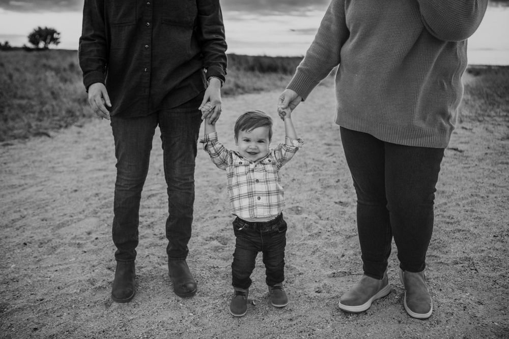 Black and white photo of small child holding hands with parents on dirt path outdoors.