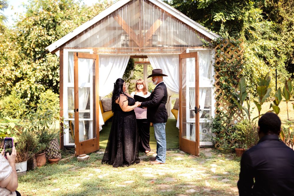 A rustic outdoor wedding ceremony takes place in front of a wooden gazebo decorated with white curtains in a garden setting.