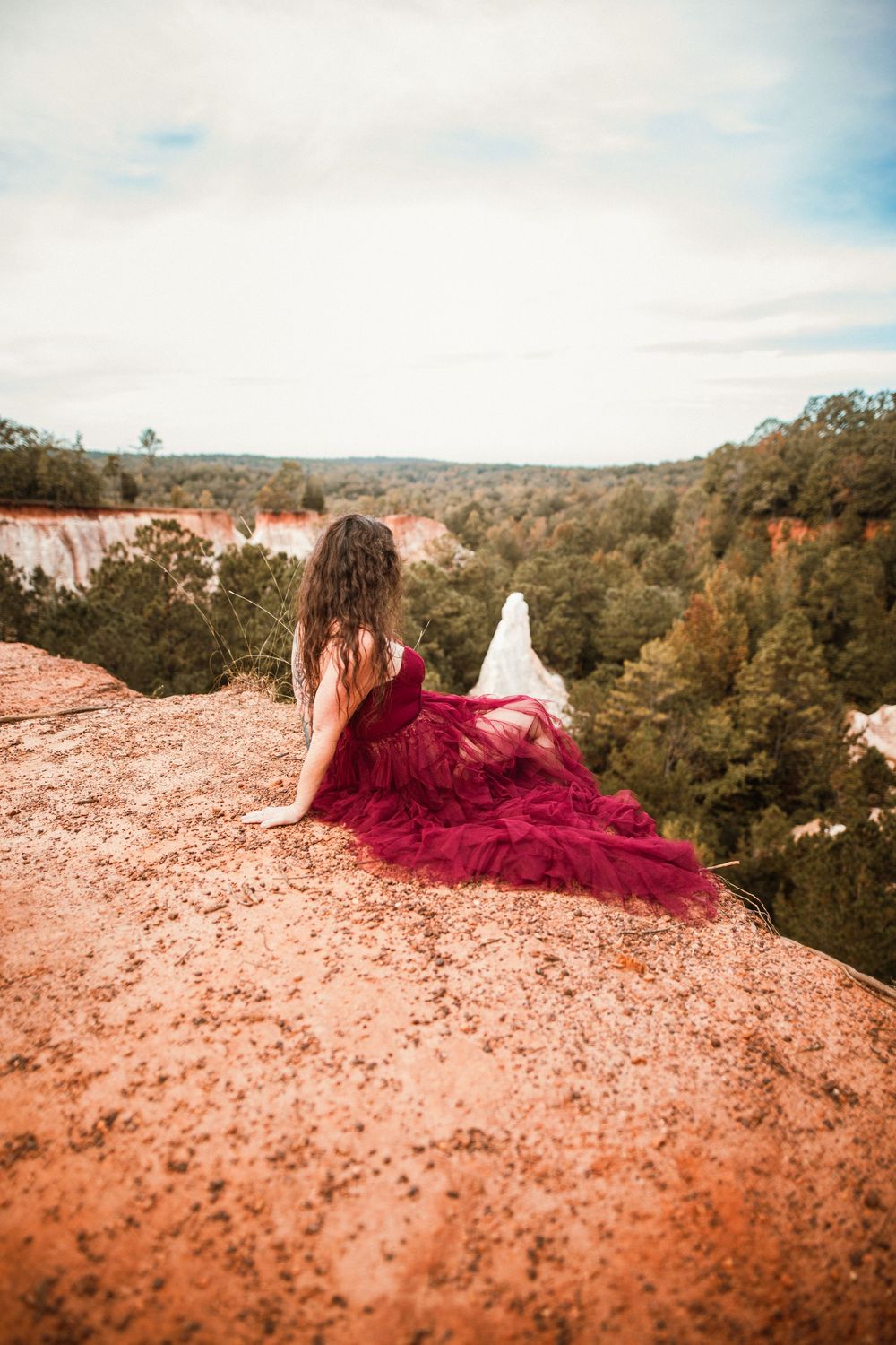 A figure in a flowing burgundy dress lies on red clay rocks overlooking a scenic forest valley landscape.