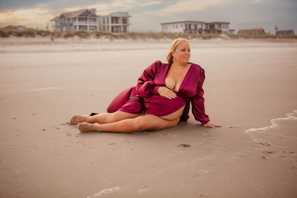 A person in a burgundy robe poses on the beach at sunset with beachfront houses visible in the background.