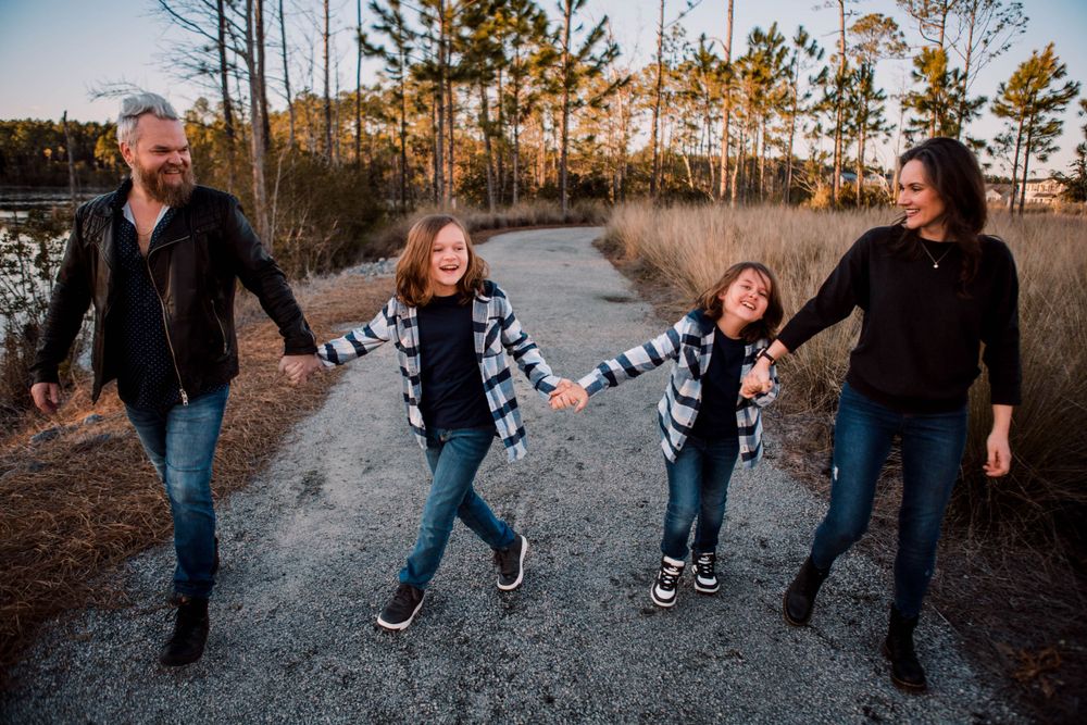 A joyful family walks hand in hand down a gravel path at sunset surrounded by pine trees.