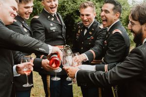 Groomsmen in black suits toasting with drinks outdoors.
