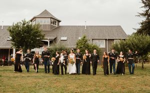 Wedding party lined up outdoors in front of rustic buildings and trees on overcast day.