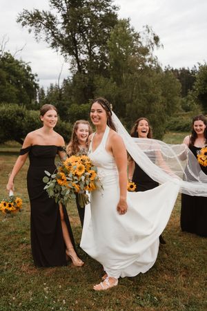 Bridesmaids in black dresses laugh with bride holding sunflower bouquets in outdoor setting.