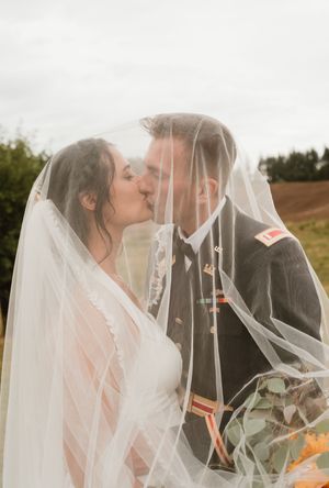 Romantic wedding couple shares intimate moment under flowing veil in natural outdoor setting.