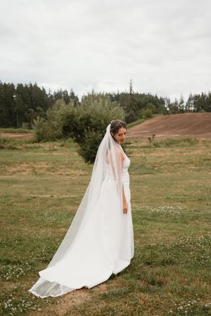 Bride's flowing white dress and veil trail behind her in rustic outdoor field setting.