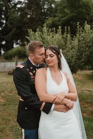 Military groom embraces bride from behind in intimate outdoor portrait.
