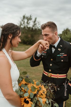 Military wedding couple shares romantic moment with sunflower bouquet in outdoor field setting.