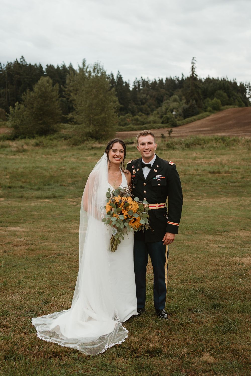 Newlyweds pose together on grassy hillside with bright sunflower bouquet and military dress uniform.