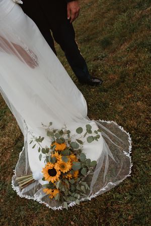 White wedding dress train with lace details and sunflower bouquet laid on grass.