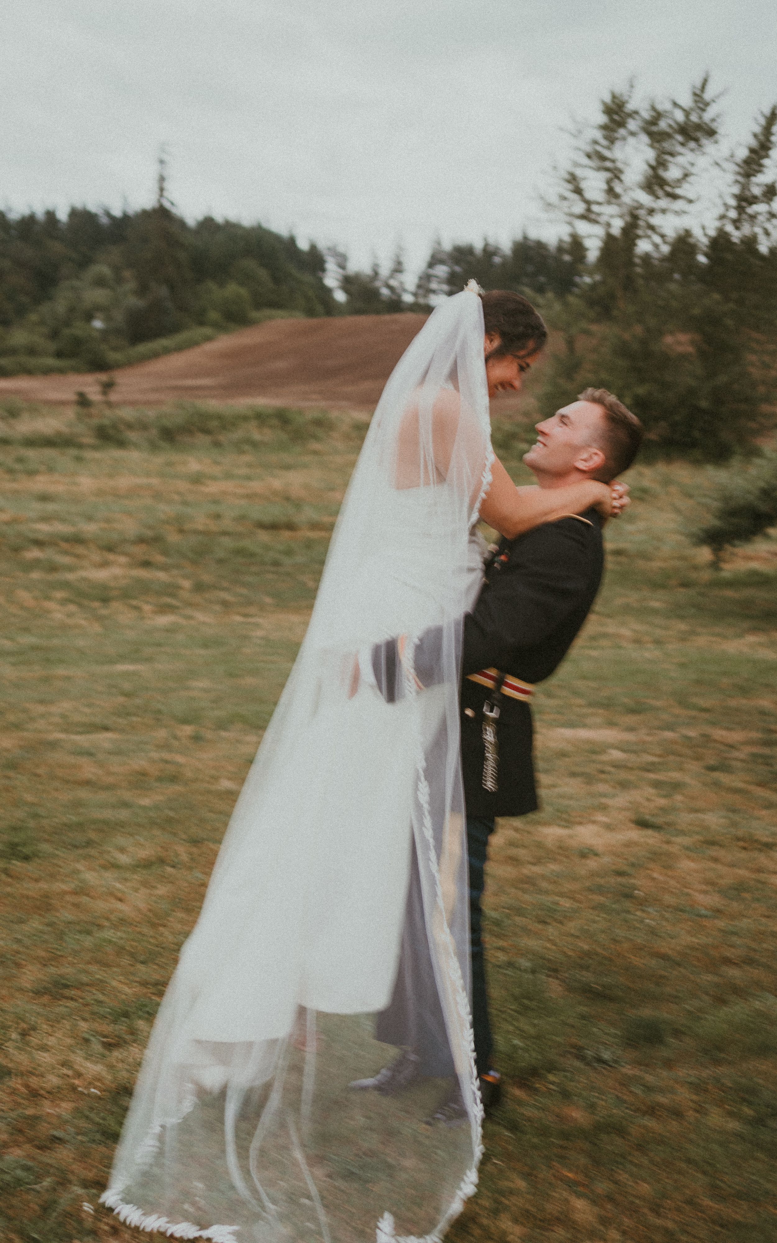 Wedding couple embraces playfully with flowing veil in outdoor meadow setting.