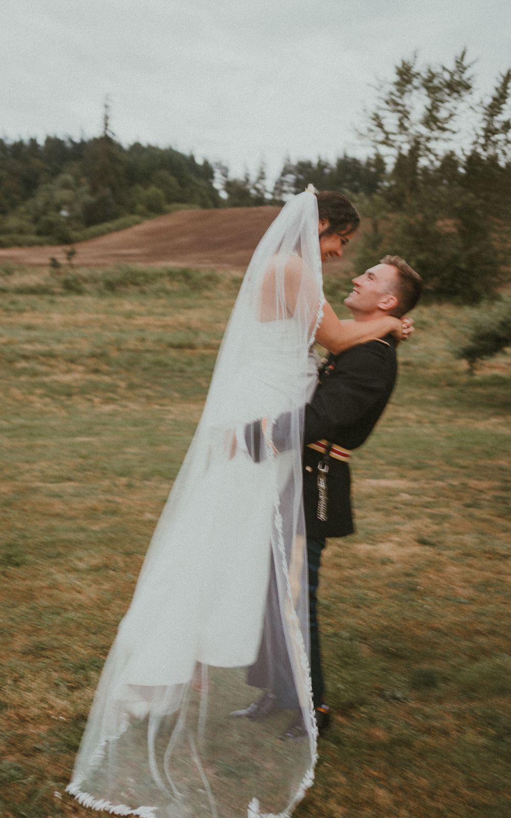 Wedding couple embraces playfully with flowing veil in outdoor meadow setting.