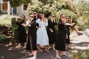 Wedding party gathers outdoors in a garden setting with bridesmaids in black dresses.