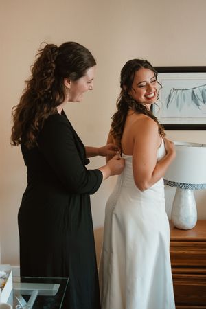 Getting ready scene shows final wedding dress preparations in a sunlit room.