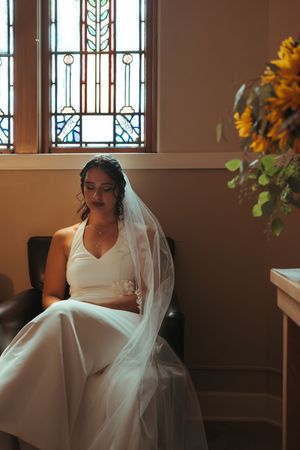 Sunlight streams through stained glass windows in church as bride sits in contemplation.