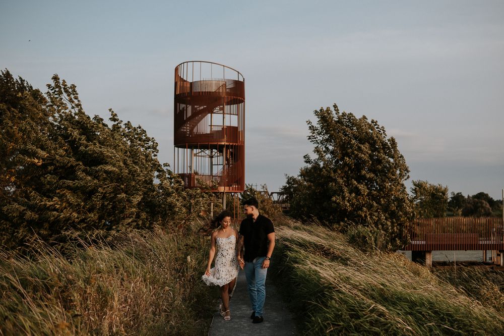 Séance couple en extérieur, deux personnes marchant dans les herbes hautes près d’une structure rustique
