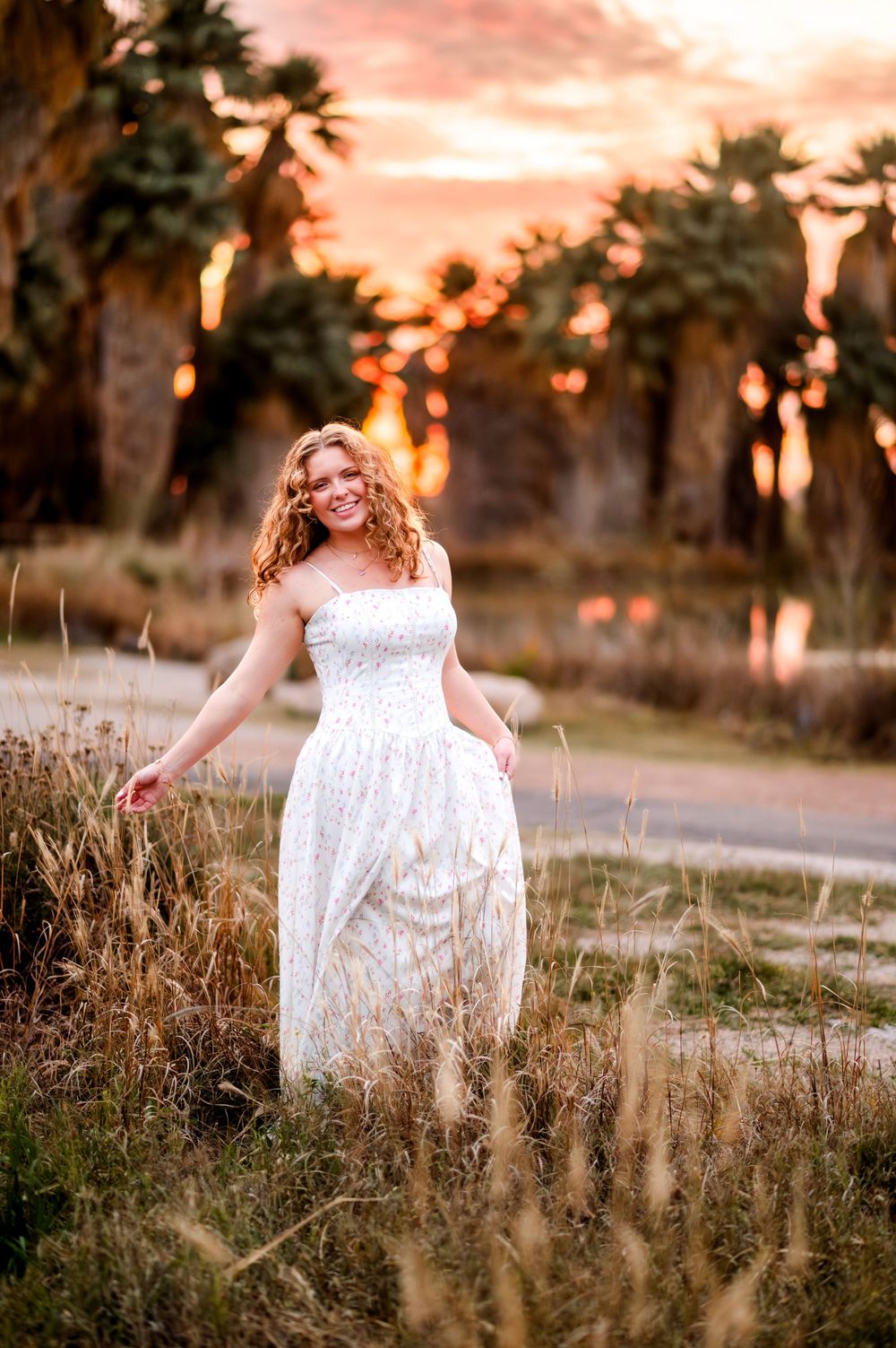 Woman in white dress standing in golden field at sunset with palm trees glowing in warm light behind her.